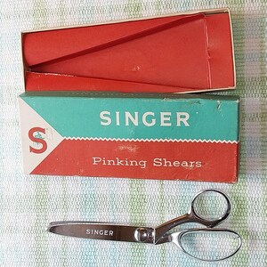 May include: Vintage Singer pinking shears with a silver blade and black handles, next to a red and teal box. The box reads "SINGER Pinking Shears" in white lettering. The box is open, revealing a red interior.