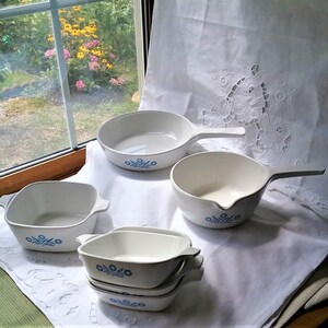 May include: A set of white ceramic cookware with blue floral designs. The set includes a square baking dish, a round casserole dish with a handle, a round skillet, and three smaller round bowls stacked on top of each other.