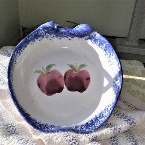 May include: A white ceramic pie dish with a blue rim and two painted red apples. The dish has a scalloped edge and a handle.