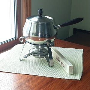 May include: A stainless steel fondue pot with a dark wooden handle and lid knob, resting on a metal stand. Next to the stand is a white box labeled "Stainless steel fondue forks" with the number 203187 printed on the side.