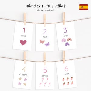 May include: A set of number flashcards, 1-6, in Spanish, hanging from a string with clothespins. Each card features a number, its Spanish word, and a corresponding illustration: a heart, butterflies, rainbows, stars, wands, and fairies.