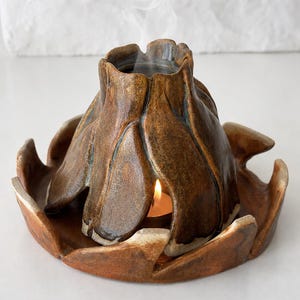 May include: A brown ceramic oil burner shaped like a blooming flower with a central opening for a tea light candle. The structure consists of a petal-like top piece resting on a matching circular base, with thin wisps of smoke rising from the center.