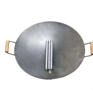 Puede incluir: Un wok grande, redondo, de color plateado con dos asas de madera. El wok est&aacute; dise&ntilde;ado para cocinar sobre fuego abierto y tiene un juego de tres patas de metal en el centro.