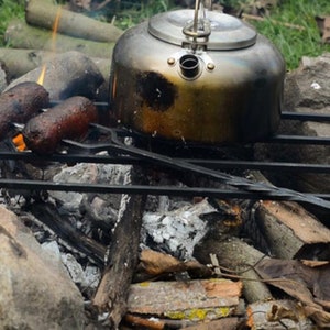 Puede incluir: Una parrilla de metal negro con dos salchichas cocinándose sobre una fogata. Una caldera negra se asienta sobre la parrilla, con llamas visibles debajo.