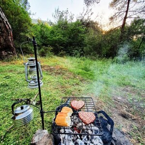 Puede incluir: Una parrilla de metal negro con dos hamburguesas en forma de corazón y dos rebanadas de pan cocinando sobre un lecho de carbón caliente. La parrilla está montada en un trípode en un área cubierta de hierba con una linterna colgando del trípode.