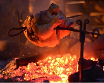 May include: A whole chicken is roasting over an open fire on a metal spit. The chicken is golden brown and has a crispy skin. The fire is made of hot coals and is glowing red.