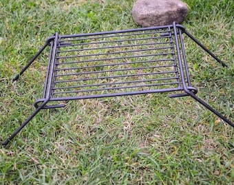 May include: Black metal grill grate with foldable legs, suitable for cooking over a campfire or open fire.