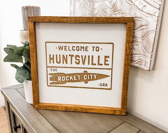 May include: A wooden sign with a white background and brown text that reads "Welcome to Huntsville, The Rocket City, USA". The sign is framed in a brown wood frame and is sitting on a gray wooden table.