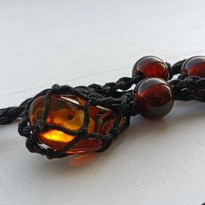 May include: A close-up of a necklace featuring a large, amber-colored stone encased in a black woven net. The necklace also includes three dark red, spherical beads. The background is a plain white surface.