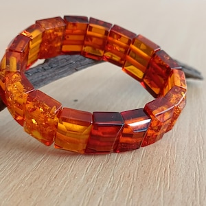 May include: A vibrant amber bracelet featuring rectangular, faceted beads in shades of orange and red. The bracelet is displayed on a piece of wood. The beads are strung together to form a circular shape.