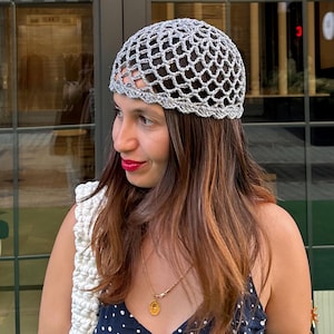 Puede incluir: Un sombrero de ganchillo gris con una corona redondeada. El sombrero lo lleva una persona que lleva un vestido azul marino con lunares blancos. Un bolso de ganchillo blanco está sobre el hombro.