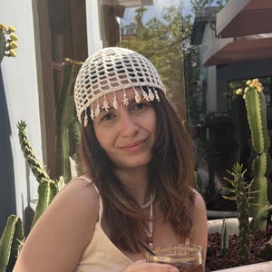 May include: A beige crochet hat with dangling embellishments. The person is wearing a beige top and skirt, holding a glass of iced coffee. Tall cacti are in the background, planted in a dark blue bathtub.