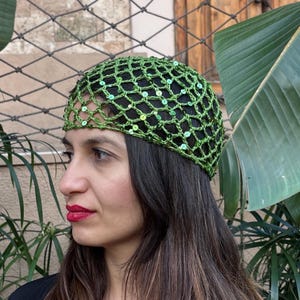 May include: A woman wearing a green crocheted head covering with sequins. The head covering is a net-like design. She is wearing a black top and has long brown hair. The background includes lush green plants and a building.