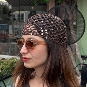 May include: A woman wearing a brown, net-style hat with sequins and brown sunglasses. She is wearing a brown top. The background includes a pub sign and greenery.