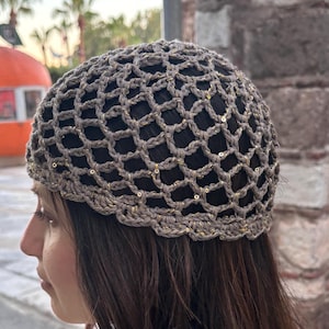 May include: A crocheted hat with a net-like design in shades of brown and gold. The hat is worn by a person with long brown hair. The background includes a brick wall, palm trees, and a round orange structure.