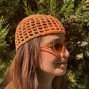 May include: An orange crochet hat with a net-like design. The hat is worn by a person wearing sunglasses and a white top. The background features green foliage.