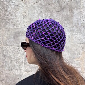 May include: A purple crochet beanie featuring a diamond-shaped open mesh pattern embellished with small, shiny purple sequins throughout. The headwear is worn over long dark hair and is paired with dark-framed sunglasses with the word Vogue on the side.