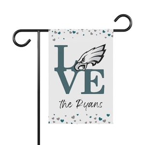 Puede incluir: Una bandera de jardín blanca con la palabra "LOVE" en letras de bloque turquesa y un logotipo de cabeza de águila. El texto "the Ryans" está debajo de la palabra "VE". Pequeños corazones turquesa y grises están esparcidos arriba y abajo.