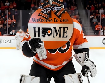 Pode incluir: Um guarda-redes de hóquei com uma camisola laranja e preta segura uma placa de madeira redonda que diz "Welcome to our HOME" com o logótipo dos Philadelphia Flyers. A placa é decorada com uma fita preta e laranja.