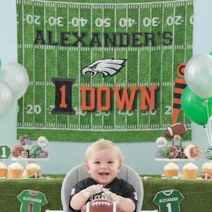 Può includere: Una festa di primo compleanno a tema calcio. Lo sfondo è un campo verde con linee e la scritta "ALEXANDER'S". Il bambino indossa una maglia nera ed è circondato da palloncini, cupcake e decorazioni a tema calcio. Il testo "1 DOWN" è visibile.