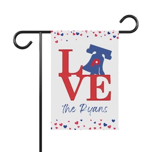 以下が含まれることがあります： 白いガーデンフラッグ。赤いブロック体の「LOVE」の文字と青いリバティベルのグラフィック。テキスト「the Ryans」は青いスクリプトです。赤と青のハートが上部と下部の端を飾っています。旗は黒い金属製のスタンドに取り付けられています。