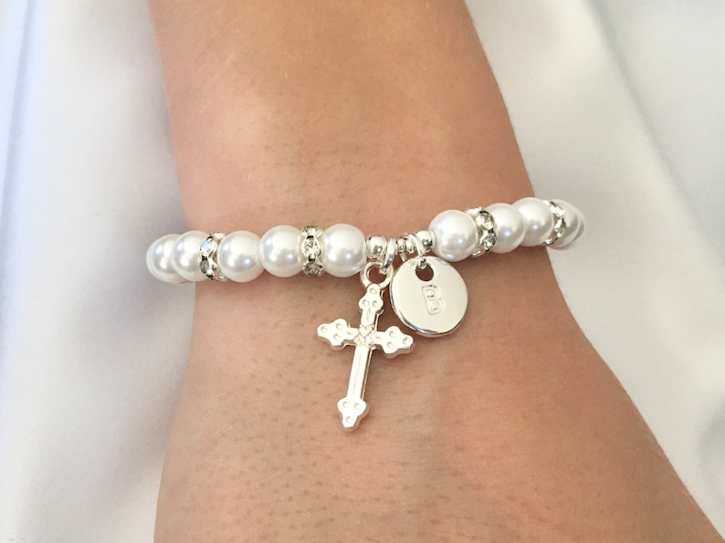May include: A white pearl bracelet with a silver cross and a silver charm with the letter "B" on it.
