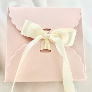 Puede incluir: Una caja de regalo rosa con un borde festoneado y una cinta de sat&eacute;n blanca atada en un lazo.
