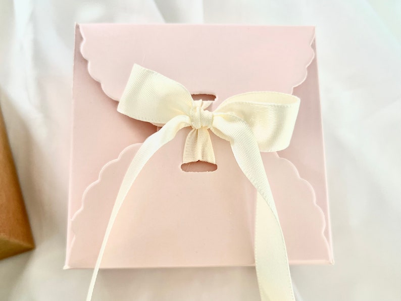 Puede incluir: Una caja de regalo rosa con un borde festoneado y una cinta de sat&eacute;n blanca atada en un lazo.