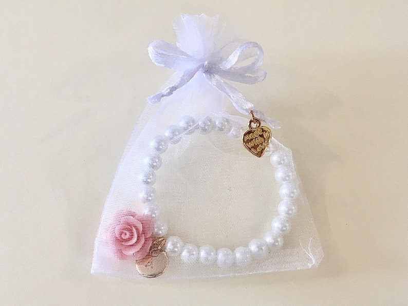May include: A white pearl bracelet with a pink flower charm and a gold heart charm. The bracelet is packaged in a white organza bag with a gold ribbon.