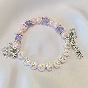 May include: A white beaded bracelet with a unicorn charm, pink and purple beads, and the name "CHARLOTTE" spelled out in gold letters. The bracelet also has a silver charm with the letters "ABC" and a ruler.