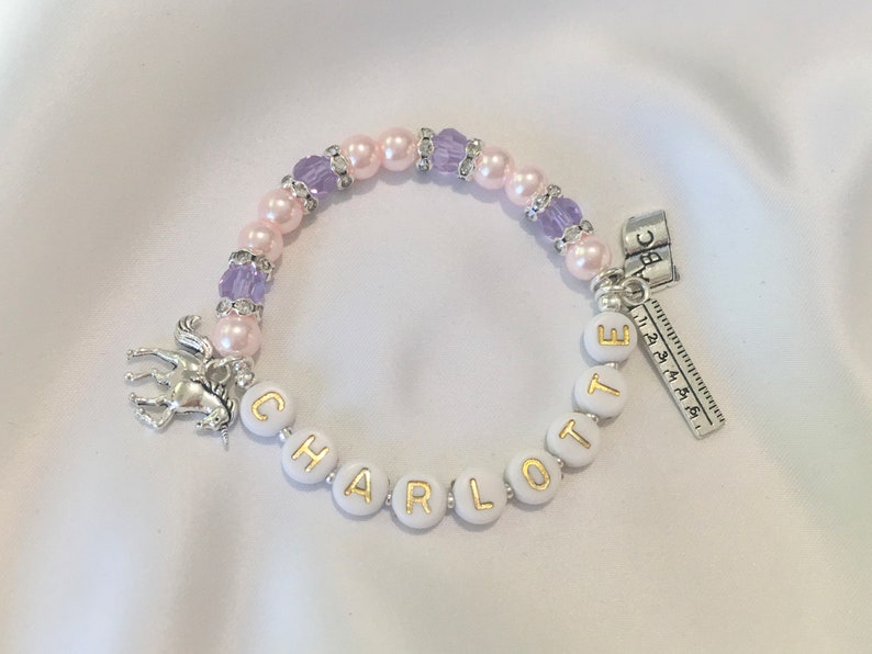 May include: A white beaded bracelet with a unicorn charm, pink and purple beads, and the name "CHARLOTTE" spelled out in gold letters. The bracelet also has a silver charm with the letters "ABC" and a ruler.