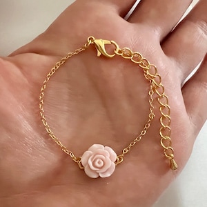 May include: A delicate gold chain bracelet with a pink flower charm. The chain is made of small, linked circles and has a lobster claw clasp.