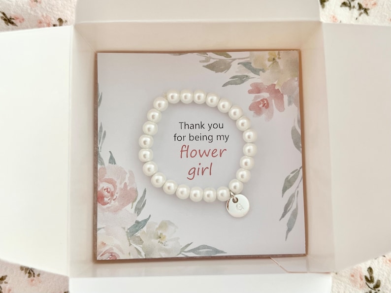 Puede incluir: Un brazalete de perlas blancas con un dije de plata que dice "Gracias por ser mi ni&ntilde;a de las flores" en una caja blanca con un dise&ntilde;o floral.