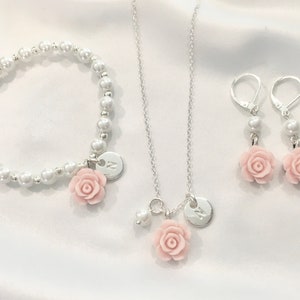 May include: A set of silver jewelry with a pearl bracelet, a necklace, and earrings. Each piece features a pink rose charm and a silver disc with the letter "N" engraved on it.