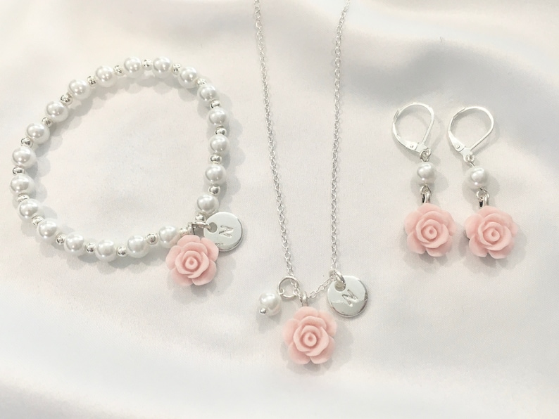 May include: A set of silver jewelry with a pearl bracelet, a necklace, and earrings. Each piece features a pink rose charm and a silver disc with the letter "N" engraved on it.