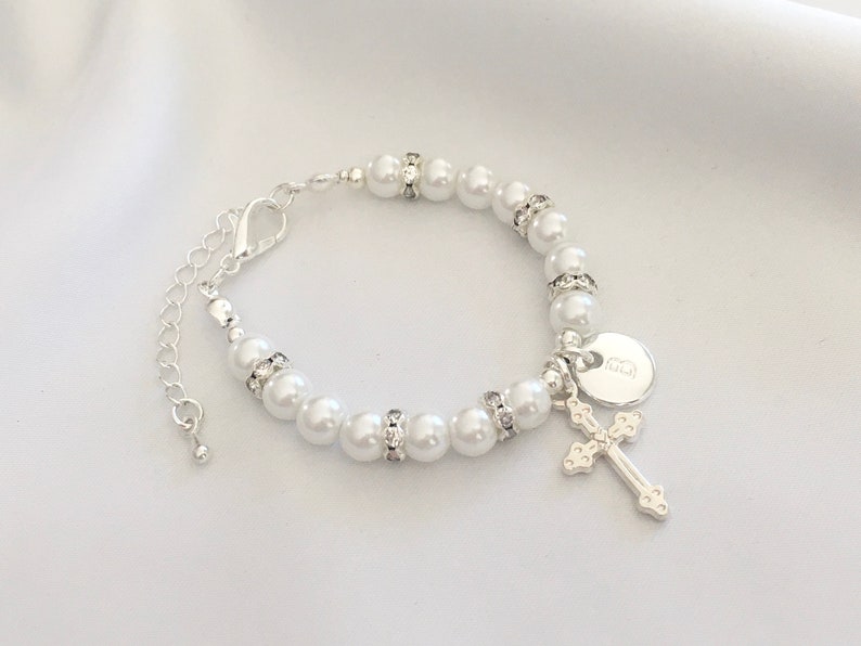 May include: A white pearl bracelet with a silver cross charm and a silver disc charm. The disc charm has the letter "E" engraved on it.
