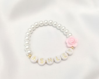 May include: A white pearl bracelet with a pink flower charm and the name "ASHLEY" spelled out in gold letters.
