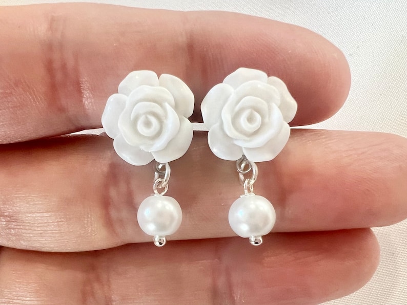 Op de afbeelding: Een paar witte rozenbloem oorbellen met een parel die aan elke bloem hangt.