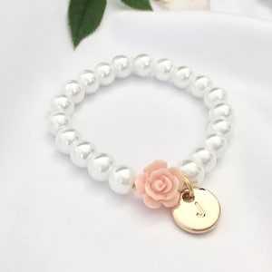 May include: A white pearl bracelet with a pink flower charm and a gold initial charm. The initial charm is a letter J.