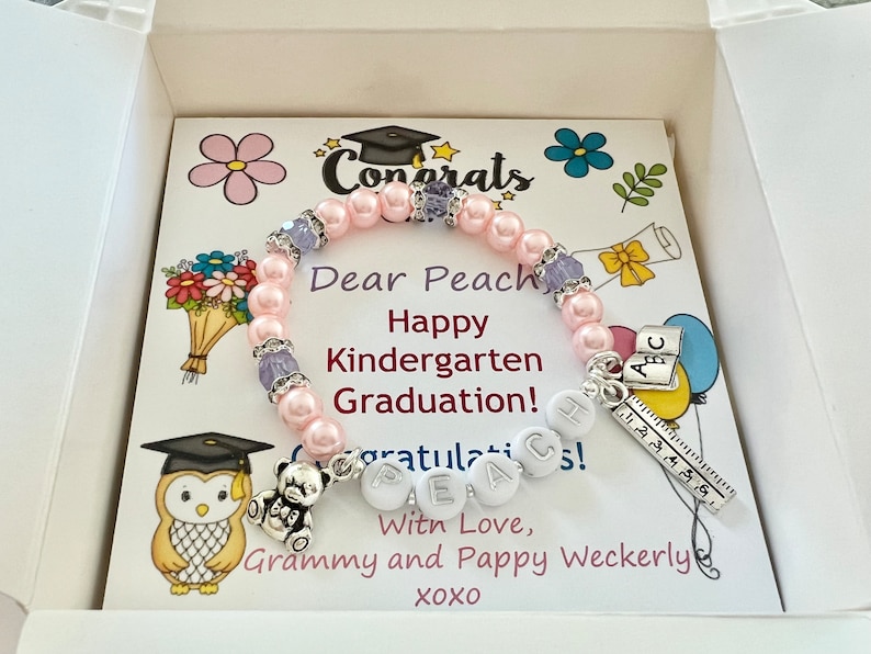 May include: A pink and purple beaded bracelet with a charm that says "Peach" and a charm that says "ABC". The bracelet is in a white gift box with a card that says "Dear Peach, Happy Kindergarten Graduation! Congratulations! With Love, Grammy and Pappy Weckerly XOXO".