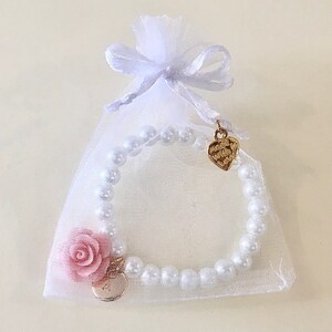 May include: A white pearl bracelet with a pink flower charm and a gold heart charm. The bracelet is packaged in a white organza bag with a gold ribbon.