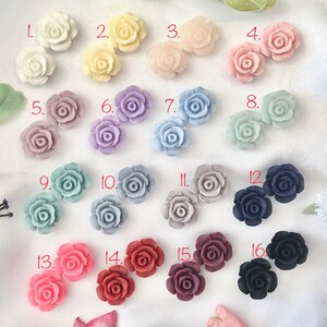 Op de afbeelding: Assortiment van 16 kleine, decoratieve, hars rozenbloem versieringen in verschillende kleuren, waaronder wit, roze, lavendel, blauw, grijs, rood en zwart. Elke roos is genummerd van 1 tot 16.