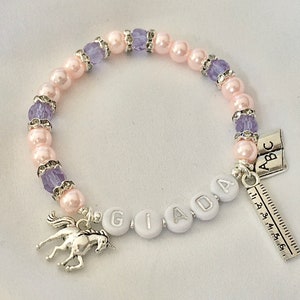 May include: A pink and purple beaded bracelet with a silver unicorn charm, a ruler charm, and a charm with the letters "ABC". The bracelet also has white beads with the letters "GIADA" on them.