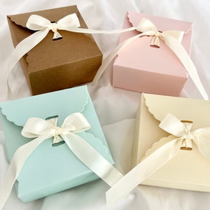Puede incluir: Cuatro cajas de regalo con bordes festoneados y lazos de cinta de sat&eacute;n. Las cajas son marrones, rosas, azul claro y crema.
