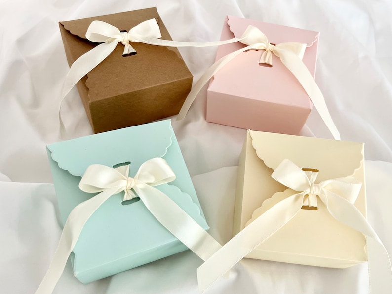 Puede incluir: Cuatro cajas de regalo con bordes festoneados y lazos de cinta de sat&eacute;n. Las cajas son marrones, rosas, azul claro y crema.