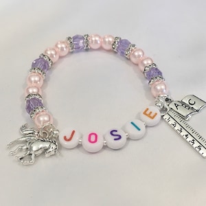 May include: A pink and purple beaded bracelet with a silver unicorn charm, a charm with the letters "JOSIE" and a charm with the letters "ABC" and a ruler.