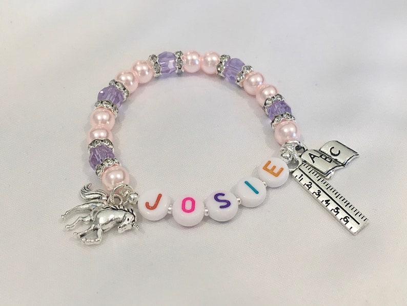 May include: A pink and purple beaded bracelet with a silver unicorn charm, a charm with the letters "JOSIE" and a charm with the letters "ABC" and a ruler.