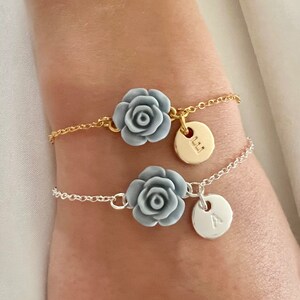 May include: Two delicate bracelets with silver and gold chains. Each bracelet features a gray rose charm and a round charm with a letter, one with the letter 'L' and the other with the letter 'A'.