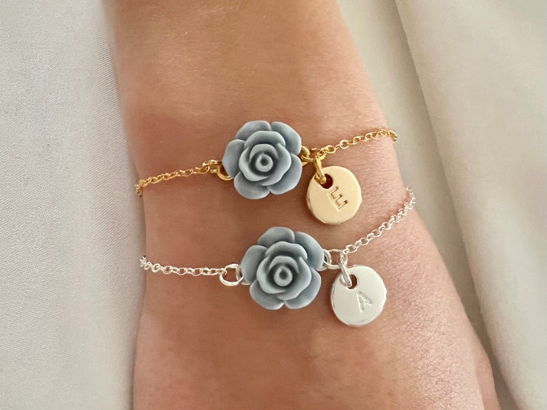 May include: Two delicate bracelets with silver and gold chains. Each bracelet features a gray rose charm and a round charm with a letter, one with the letter 'L' and the other with the letter 'A'.