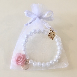 May include: A white pearl bracelet with a pink flower charm and a gold heart charm. The bracelet is packaged in a white organza bag with a white satin ribbon.
